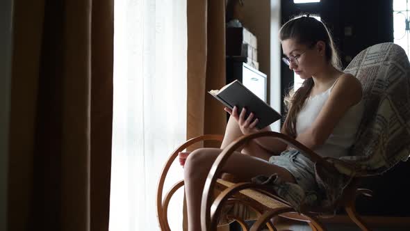 woman reads a book sitting in a rocking chair in front of the window alt