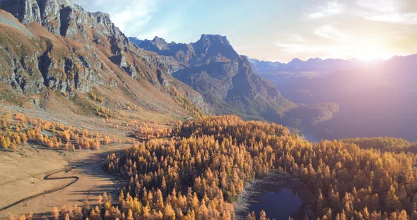 Forward Aerial Over Alpine Mountain Valley Lake and Orange Larch Forest Woods in Sunny Autumn alt
