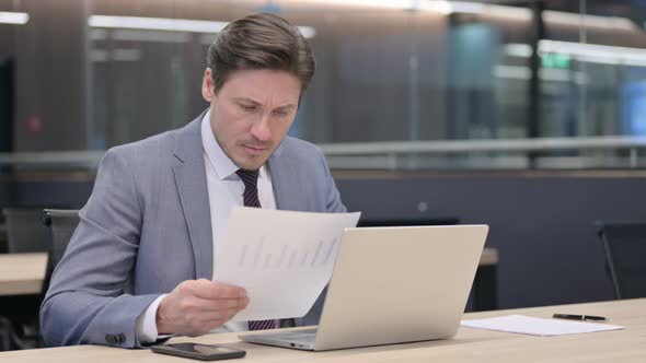 Middle Aged Businessman having Loss on Documents while using Laptop  alt