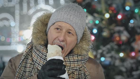 A Hungry Elderly Man Eats in a Food Court at a Christmas Market on the Street in Winter