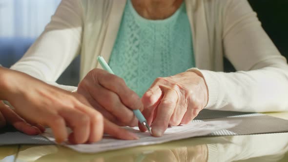 Elderly Woman in Glasses Signing Document with Help of Volunteer 