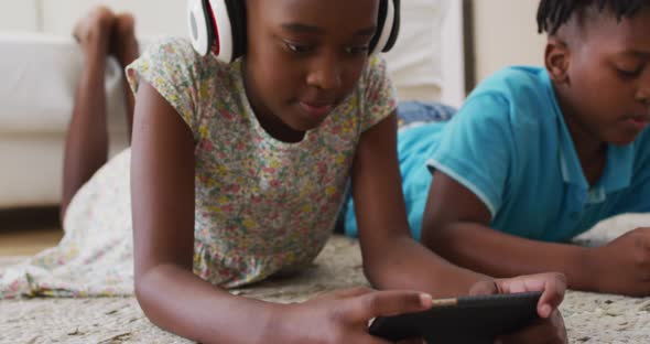 African american brother and sister using electronic devices lying on the floor at home alt