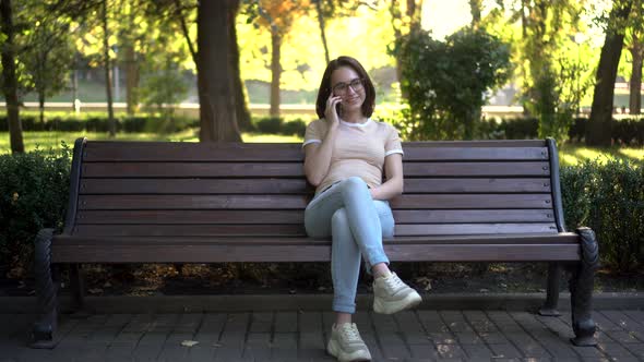 A Young Woman in Glasses Sits on a Bench Speaks on the Phone. In the Background Nature alt