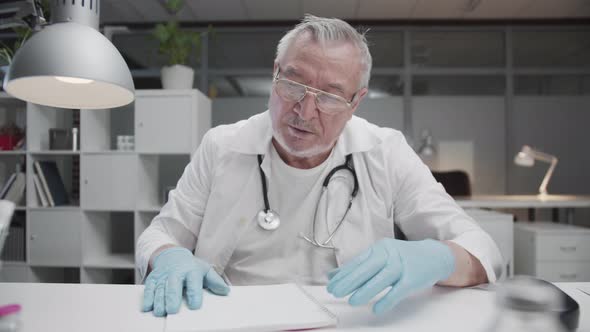 A Tired Elderly Experienced Doctor in Latex Gloves and Glasses After Finishing a Shift at the alt