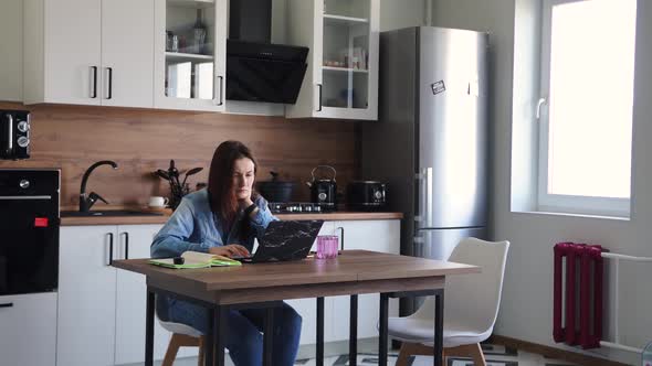 Woman Working at Laptop While Sitting at the Table in the Kitchen at Home