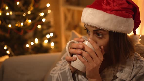 Woman in Pajama and Red Santa Hat Sitting on Couch Watching TV and Drinking Hot Chocolate From Mug alt
