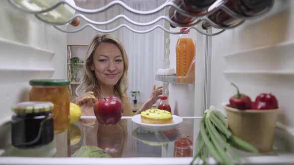 Beautiful Young Woman Opens the Refrigerator Door Looks Inside Takes an Apple and Then Happily Takes alt