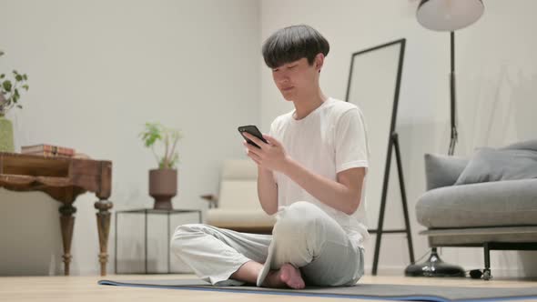 Young Asian Man Using Smartphone on Yoga Mat at Home alt