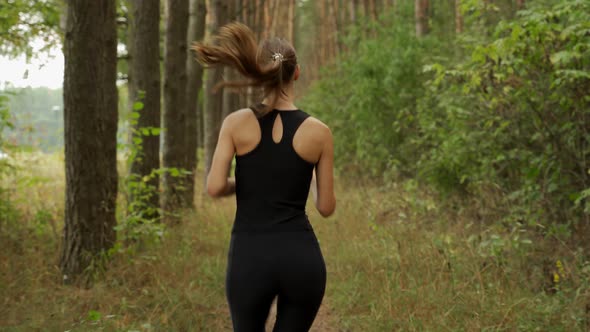Rear View of a Young Woman Running Through a Pine Forest on a Sunny Morning alt
