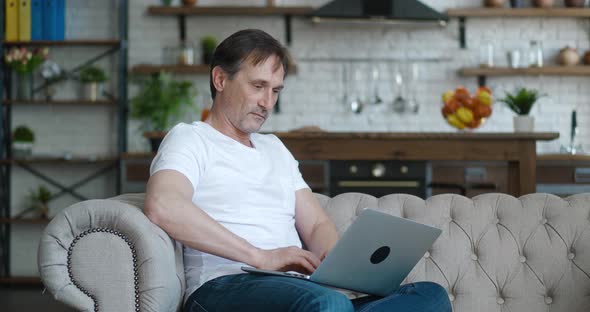 Senior Mature Man Typing on Laptop Browsing Internet Using Apps Sit on Sofa Alone at Home Apartment alt