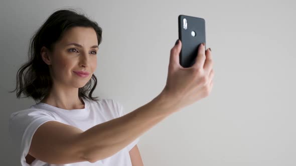 Woman in a White T-shirt on a Light Wall Makes a Selfie alt