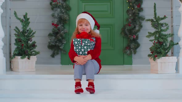Cheerful Smiling Toddler Child Girl Kid Sitting at Decorated House Porch Holding One Christmas Box alt