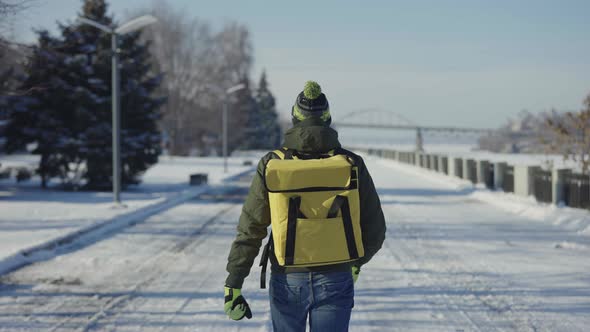 Back View of Courier with Large Yellow Thermo Bag Walking Along Snow Covered City Street alt