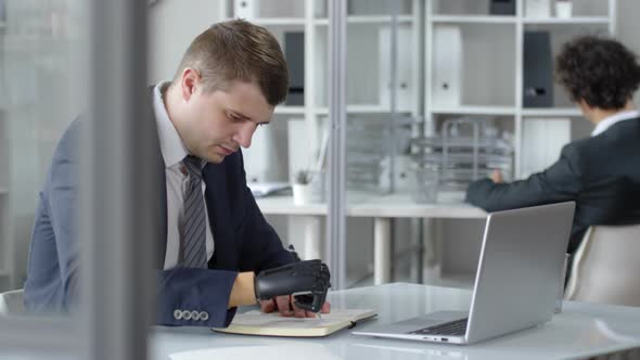 Businessman with Prosthetic Hand Writing in Notepad at Office Desk alt