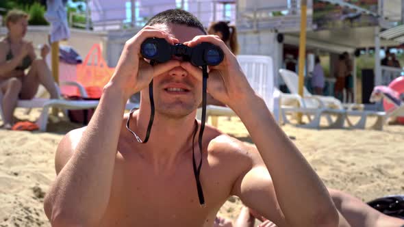 Man on the Beach in Swimming Trunks is Watching Someone Through Binoculars alt