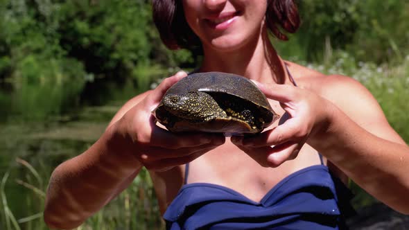 Woman Holds Funny Turtle in Arm and Smiles on River with Green Vegetation alt