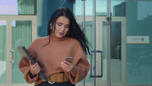 Young Elegant Serious Successful Business Woman Walks on Background Office Building Holding Document alt