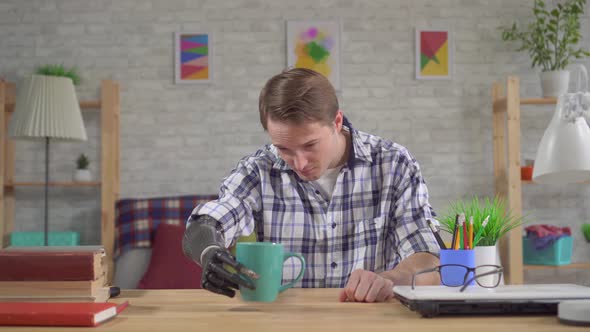 Young Man Sitting at a Table Takes a Cup with a Bionic Prosthetic Hand alt