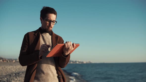 Bearded Man Is Using Flatbed Computer Sitting on Seashore with Panorama of City alt
