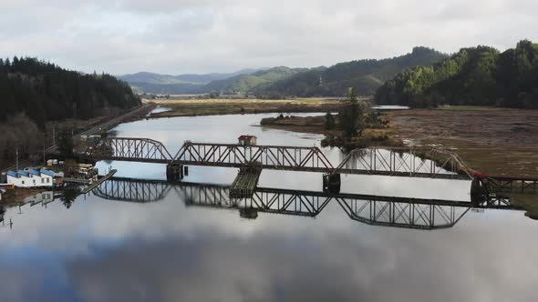 wide Siuslaw river passes under steel railbridge structure. Cushman, OR, USA. push in aerial alt