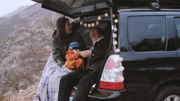 Family  sitting in the open trunk of a black car with baby against the backdrop of autumn mountains alt