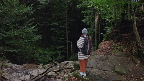 Red Head Girl Tourist with a Backpack, Stands on the Edge of a Cliff of a Waterfall Against a Foggy alt