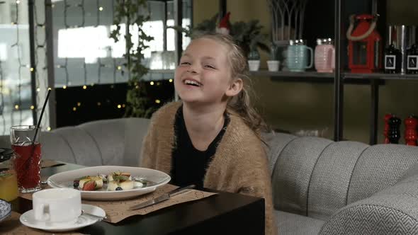 a Little Girl with Blonde Hair Laughs Merrily Sitting in a Restaurant