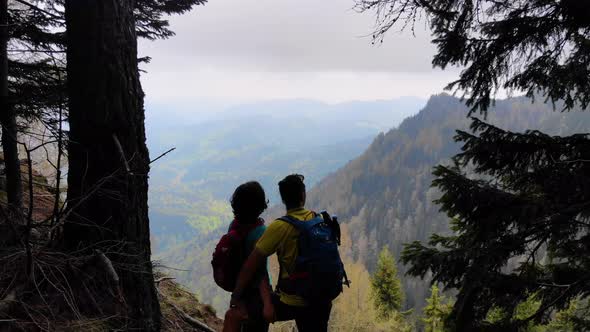 Aerial Flight Over Couple Sitting on Cliff Edge. Young couple hiking on top of the mountai. alt