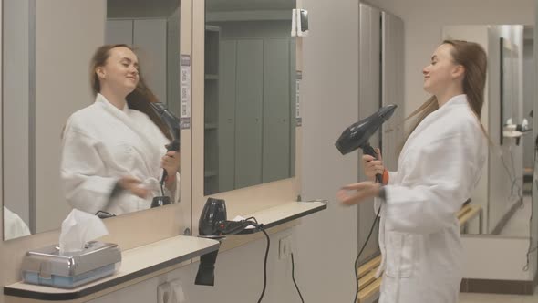 Young Charming Sportswoman in White Bathrobe Drying Hair with Dryer in Gym Locker Room. Confident alt