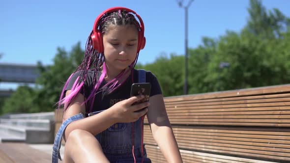 Girl Listening to Music on Headphones Using Smartphone Sitting on a Bench with a Good Mood in Summer alt