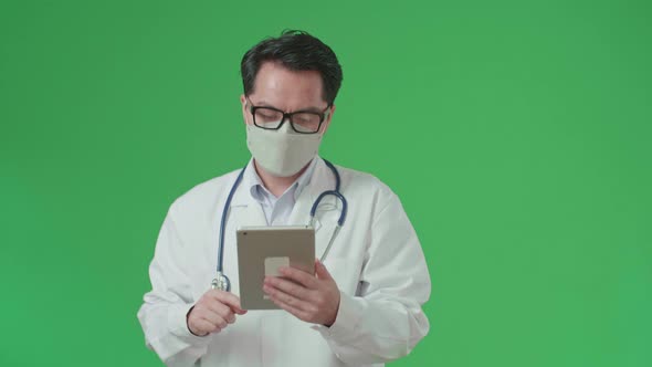 Asian Doctor With Stethoscope Wearing Mask, Using Tablet While Walking On Green Screen alt