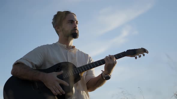 Spanish Bearded Man Playing Guitar Outdoors Against a Blue Sky Lit with Setting Sunlight alt