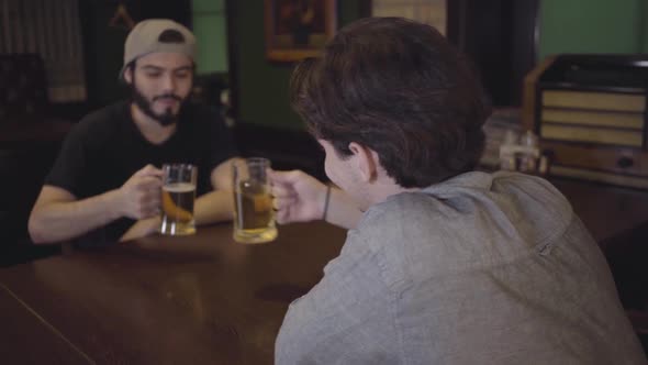 Two Guys Drink Beer Clinking Glasses While Sitting at a Table in a Pub alt