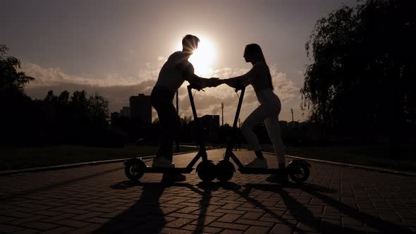 Silhouette of a Young Couple in Love on Electric Scooters in the Park at Sunset alt