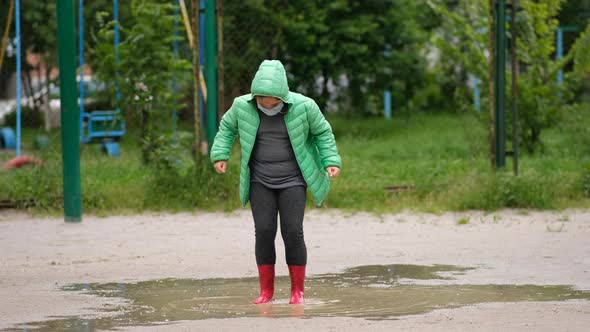 A little girl in a medical mask and rubber boots jumps through puddles on a spring day after rain. 
