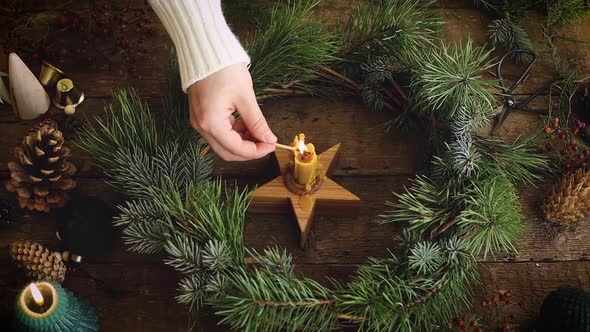 Hand in sweater lighting up candle in christmas wreath on rustic wooden table alt