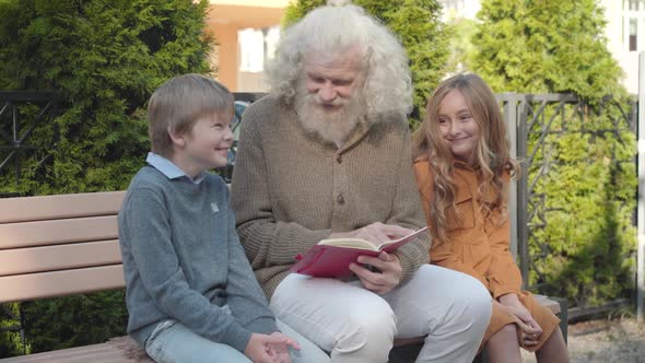 Positive Grandfather Sitting with Grandchildren on Bench and Reading Book. Portrait of Happy Relaxed alt