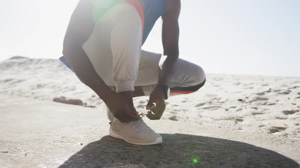Midsection of african american man tying shoelaces during exercise outdoors on beach alt