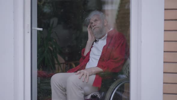 Thoughtful Sad Disabled Senior Man in Wheelchair Sitting Behind Glass Door Looking Out on Rainy Day alt