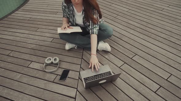 Caucasian Redhaired Girl Sits on a Wooden Floor Makes Plans Using a Notebook and a Computer