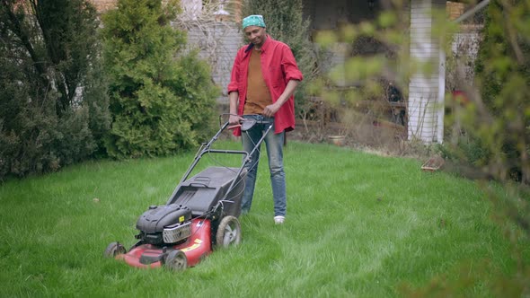 African American Young Man Scratching Head Standing on Green Backyard Lawn with Lawn Mower alt