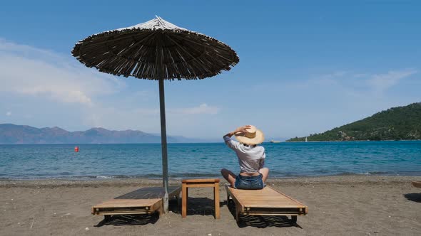 Girl in Shirt and Hat Sitting on the Beach on a Sunbed Under an Umbrella alt