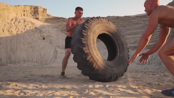 Two Athletes Train in Active Mode on the Beach Doing push-UPS and Pushing a Huge Wheel alt