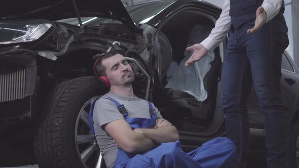 Exhausted Caucasian Young Man in Earphones Sleeping Leaning Against Car Wheel As Unrecognizable Guy alt