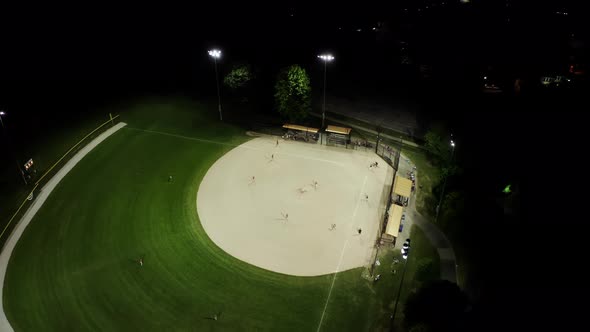 Aerial Drone Footage Of Baseball Field At Night With People Playing Baseball On Grass Field alt