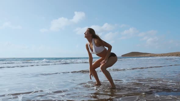 Girl in Sportswear Shorts and T-shirt Performs Jumps with Squats on the Beach Near the Ocean alt