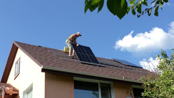 Man Instals Solar Panel on on Bituminous Tiles Roof of Small Countryside House