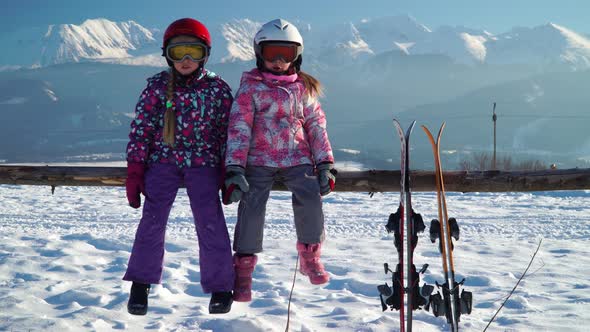 Little Girls in Outwear and Masks with Helmet Sitting on Wood Fence with Skis Near Against Mountains