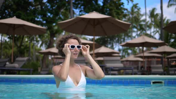 Caucasian Woman in a White Swimsuit and Wears Her Sunglasses While Standing in a Swimming Pool alt