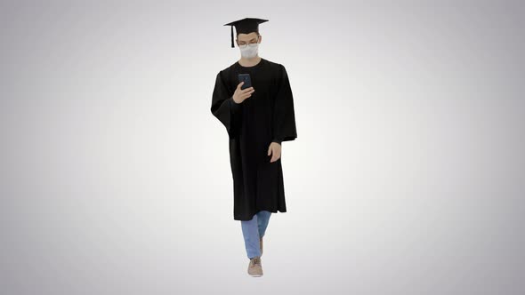 Young Man with Graduation Gown Walking in Medical Mask Using Smartphone on Gradient Background alt